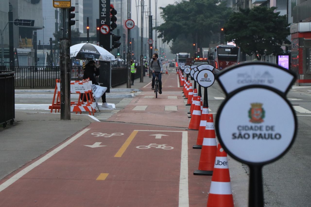 Em primeiro plano, à direita da foto, totens redondos com o logotipo da Prefeitura de São Paulo e da "ciclolazer sp", e ao fundo da foto está a ciclofaixa de lazer da Avenida Paulista.